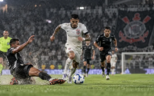 Jogador do Corinthians de uniforme branco conduz a bola em gramado molhado enquanto um adversário tenta o carrinho