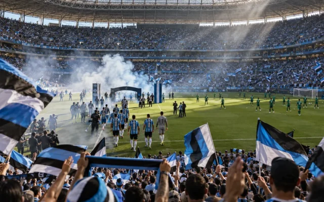 Arena do Grêmio lotada, com bandeiras tricolores, jogadores entrando entre fumaça e Palmeiras aquecendo sob a luz dramática da manhã